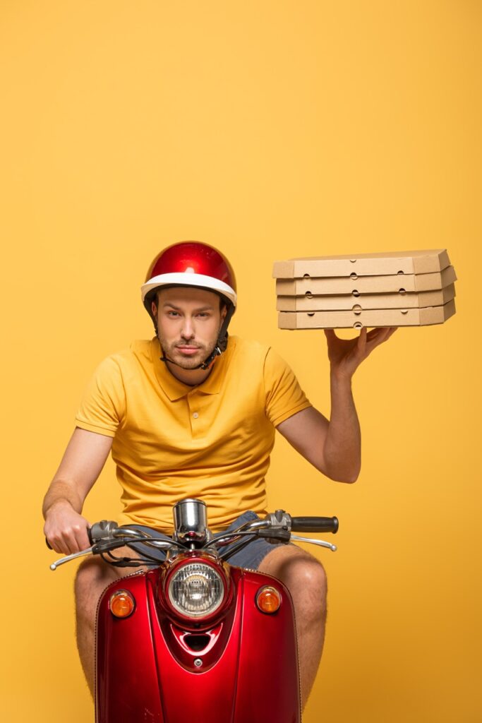 concentrated delivery man in yellow uniform riding scooter with pizza boxes isolated on yellow
